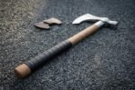 Bearded axe with knotwork engraved steel head and black wrapped ash haft lying on gravel beside a leather edge guard