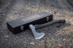 Bearded axe with knotwork panel on the head and dark hardwood haft lying in front of its black engraved wooden case on gravel