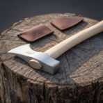 Bearded hand axe with wedged ash handle and struck rune marks lying on a tree stump next to two brown leather covers