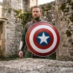 Bearded man holding a painted round steel shield with concentric bands and a star in front of a ruined stone wall