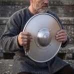 Bearded man holding a round steel buckler with an engraved Mjolnir boss and rolled rim in front of him