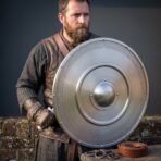 Bearded man in mail and a leather jerkin holding a steel round shield with domed boss and knotwork band at his side