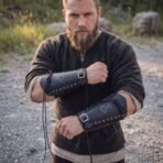 Bearded man wearing black leather bracers with Mjolnir engraving and laced eyelets, arms crossed outdoors