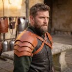 Bearded man wearing tan leather pauldrons with layered lames and an engraved wolf head cap in an armour courtyard