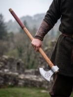 Bearded Viking axe with Vegvisir engraved head and long hardwood haft carried in one hand near ruined stone walls