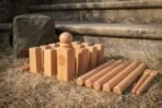 Beech kubb king with an engraved vegvisir standing among the blocks and throwing batons on dry grass, canvas bag behind