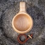 Birch kuksa wooden cup seen from above on a granite rock with a leather carry cord and brass fitting coiled beside it