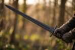 Black finished steel sword with fuller held in both gloved hands, blade angled up against a sunlit woodland
