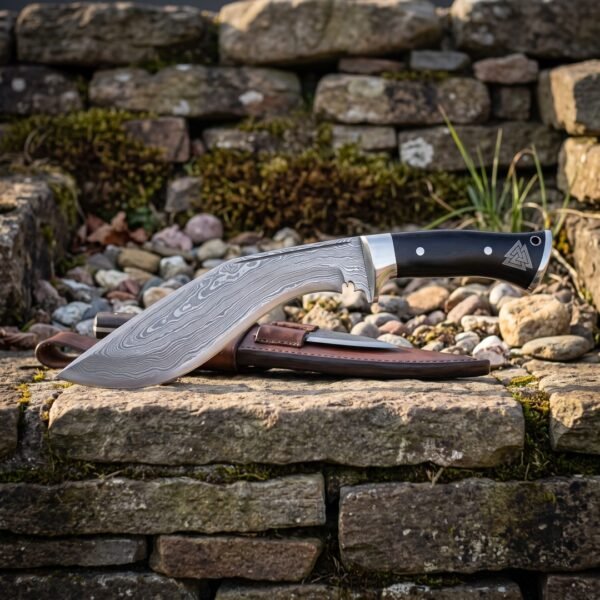 Black handle damascus kukri with a polished steel bolster and engraved valknut butt cap resting on a dry stone wall beside its brown leather sheath and companion blades.