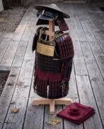 Black lacquered samurai armour with crimson lacing and brass rune plaque on a wooden stand beside a folded red cloth