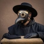 Black leather plague doctor mask with brass lens rims worn with a wide brimmed black hat and black gloves, seated at a stone table