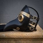 Black leather plague doctor mask with brass rimmed lenses and an engraved brass brow band, laid on a stone plinth with three buckle straps showing
