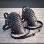Pair of black leather scale pauldrons with riveted overlapping scales, valknut stamped panels and buckled straps on a grey wooden table