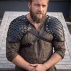Black leather scale pauldrons with silver rivets and crossed black straps worn over a linen tunic by a bearded man
