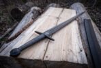 Close up of a black oxide arming sword hilt with wire wrapped grip, disc pommel and valknut mark on a split log, scabbard behind