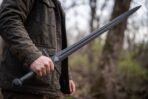 Black oxide arming sword with valknut marked fullered blade held at the side by a man in a brown jacket in bare woodland