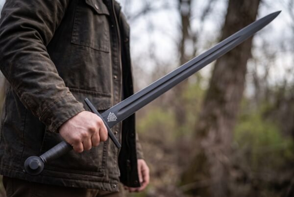 Black oxide arming sword with valknut marked fullered blade held at the side by a man in a brown jacket in bare woodland