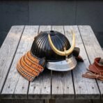 Black ribbed kabuto helmet with a brass crescent crest and knotwork brow band sitting on a weathered wooden table