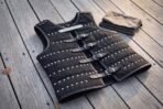 Black riveted leather brigandine body armour laid at an angle on grey wooden decking, four steel buckles down the front and a folded grey cloth