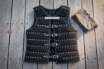 Black riveted leather brigandine body armour with four steel buckle straps and an engraved rune plaque, laid flat on grey weathered decking boards outdoors.