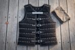 Black riveted leather brigandine laid flat on wooden decking, rows of steel rivets, four buckled straps and a rune cut plaque at the chest