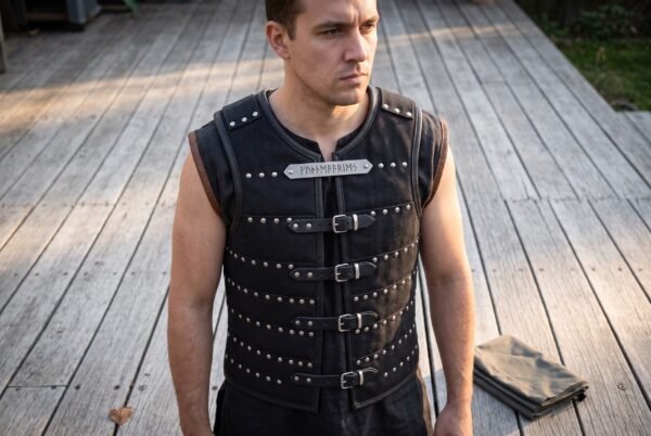 Black riveted leather brigandine with four buckled straps and a rune cut chest plaque worn over a black shirt by a dark haired man on decking