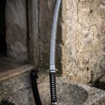Black saya wakizashi with a folded Damascus blade and cord wrapped handle standing on a weathered stone trough in front of a rendered farmhouse wall