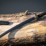 Black wakizashi short sword in blackened 1095 steel with a cord wrapped tsuka and dark tsuba resting on a fallen bleached log beside its matte black saya
