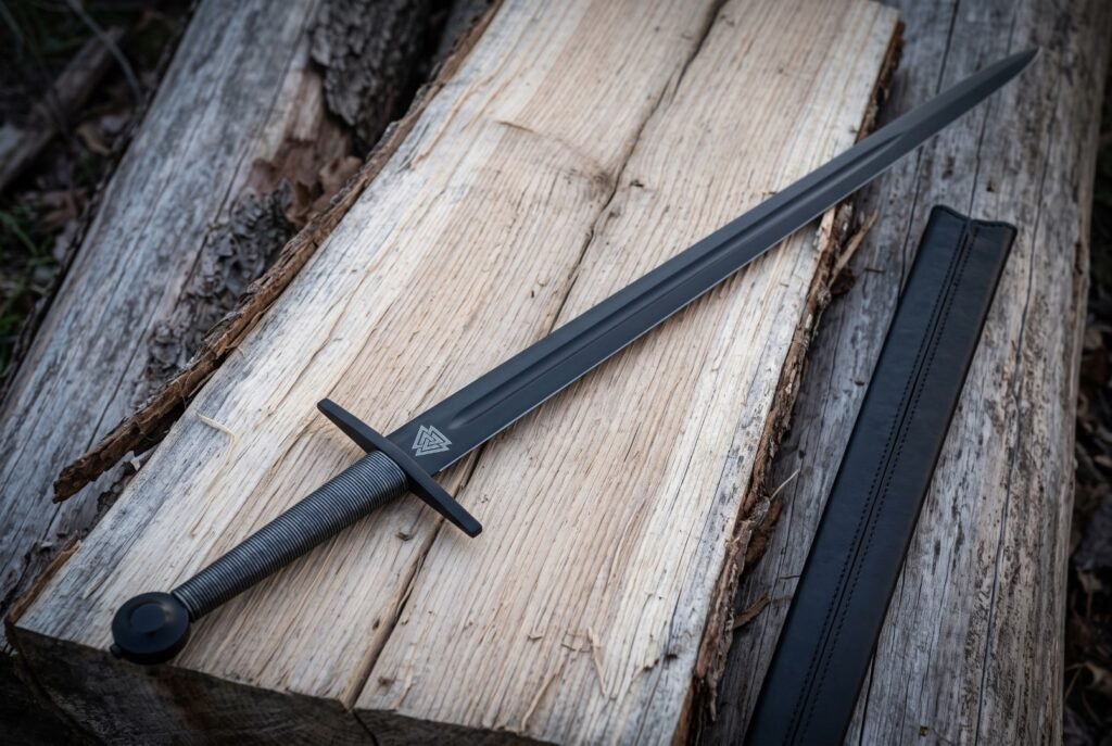 Blackened 1095 steel valknut arming sword with a wire wrapped grip and black leather scabbard resting on weathered pale timber planks in daylight