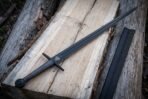 Blackened 1095 steel valknut arming sword with a wire wrapped grip and black leather scabbard resting on weathered pale timber planks in daylight