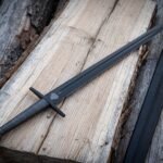 Blackened 1095 steel valknut arming sword with a wire wrapped grip and black leather scabbard resting on weathered pale timber planks in daylight