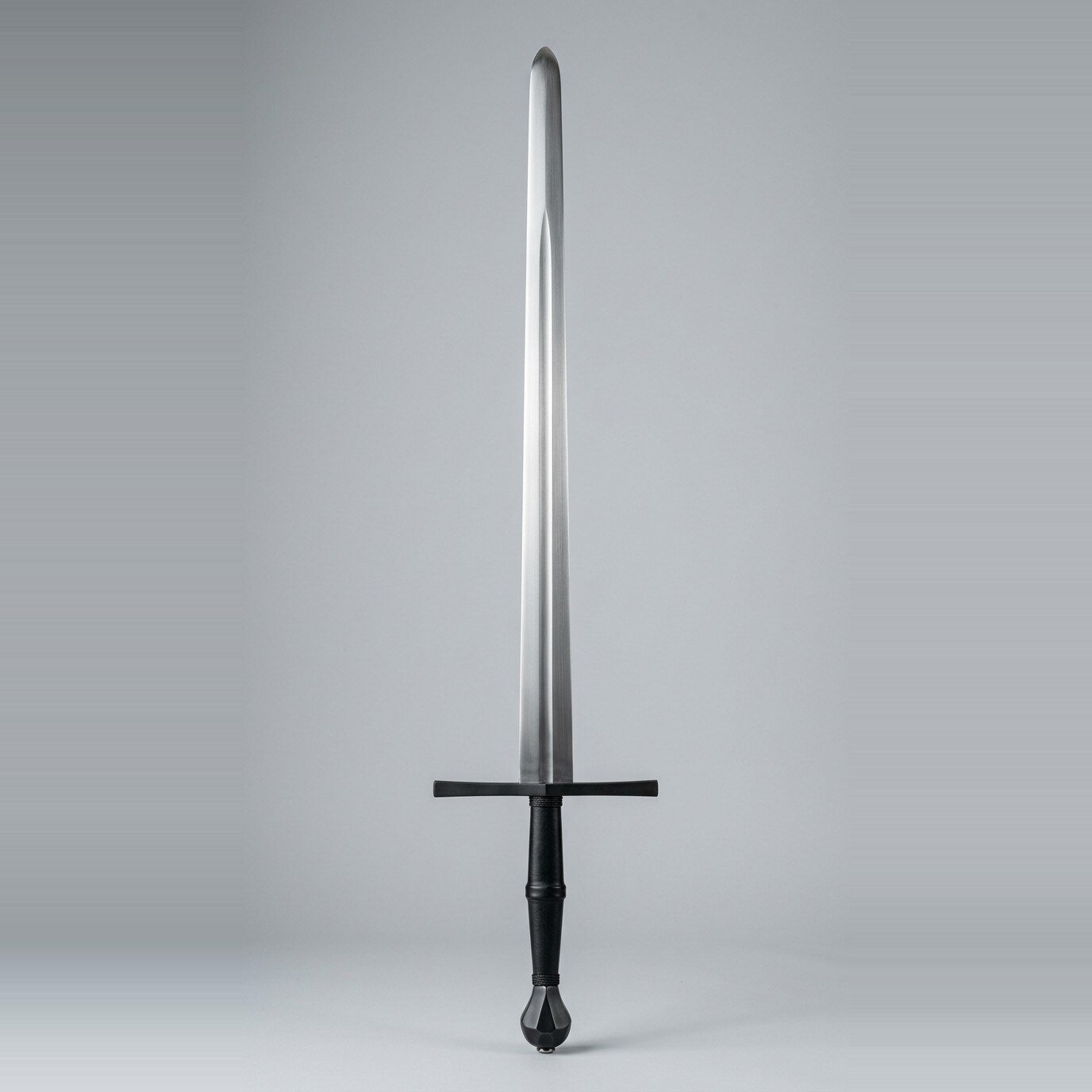 Blackened blunt longsword trainer Blackened blunt longsword trainer with a dark cross and pommel, black leather grip and a fuller running two thirds of the blade shown against a plain studio backdrop