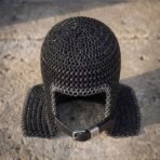 Blackened butted chainmail coif seen from above on a stone slab, showing the bright ring face course and black leather chin strap with buckle