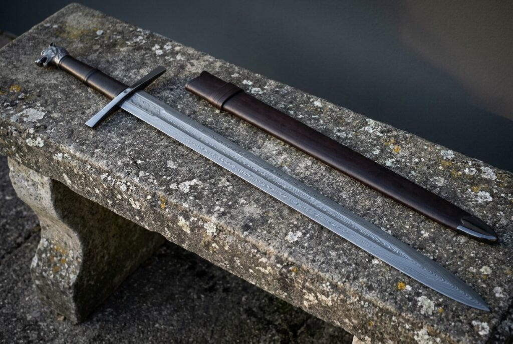 Blackened cast wolf head pommel sword in pattern welded Damascus resting on a lichen covered stone bench next to its dark brown leather scabbard in daylight