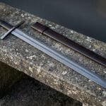 Blackened cast wolf head pommel sword in pattern welded Damascus resting on a lichen covered stone bench next to its dark brown leather scabbard in daylight
