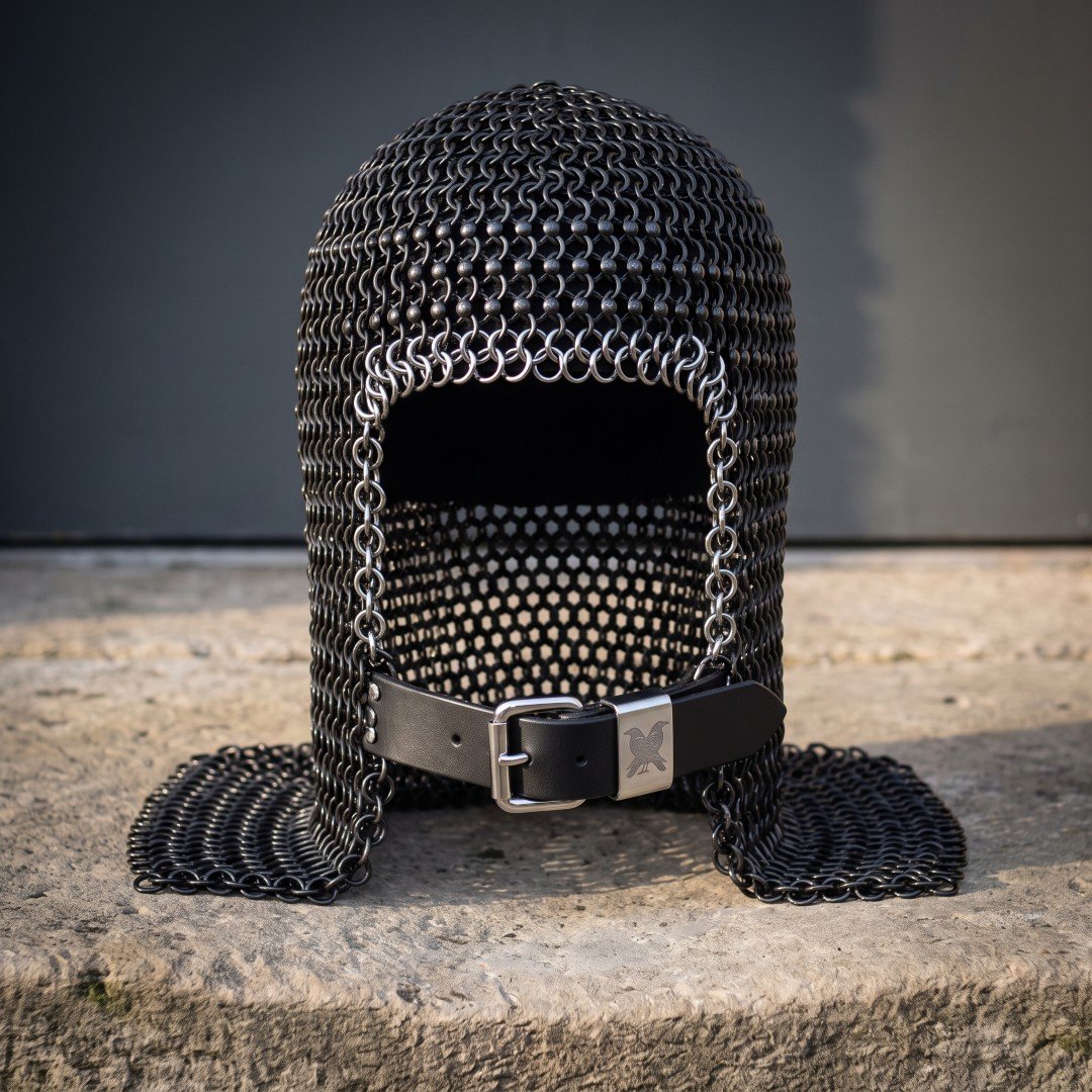 Blackened chainmail armor coif in butted mild steel mail with an integral ventail face flap, bright ring face course and Blackened chainmail armor coif in butted mild steel mail with an integral ventail face flap, bright ring face course and raven stamped leather strap on a stone ledge
