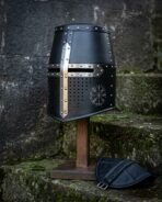 Blackened crusader helmet, a hand forged 16 gauge great helm with a polished steel cross reinforce, domed rivets and a chased Vegvisir, on a wooden stand by mossy steps