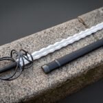 Blackened flamberge sword with a polished wavy 1095 blade and swept ring hilt lying on a granite ledge beside its black leather scabbard with steel fittings.