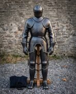 Blackened hand forged plate armour suit on a wooden stand against a stone wall, showing fluted breastplate, engraved runes, articulated pauldrons, gauntlets and sabatons.