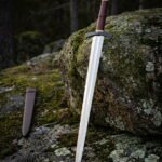 Blackened knotwork pommel viking arming sword in hand forged 1095 steel with an oxblood leather grip standing against a lichen covered forest boulder