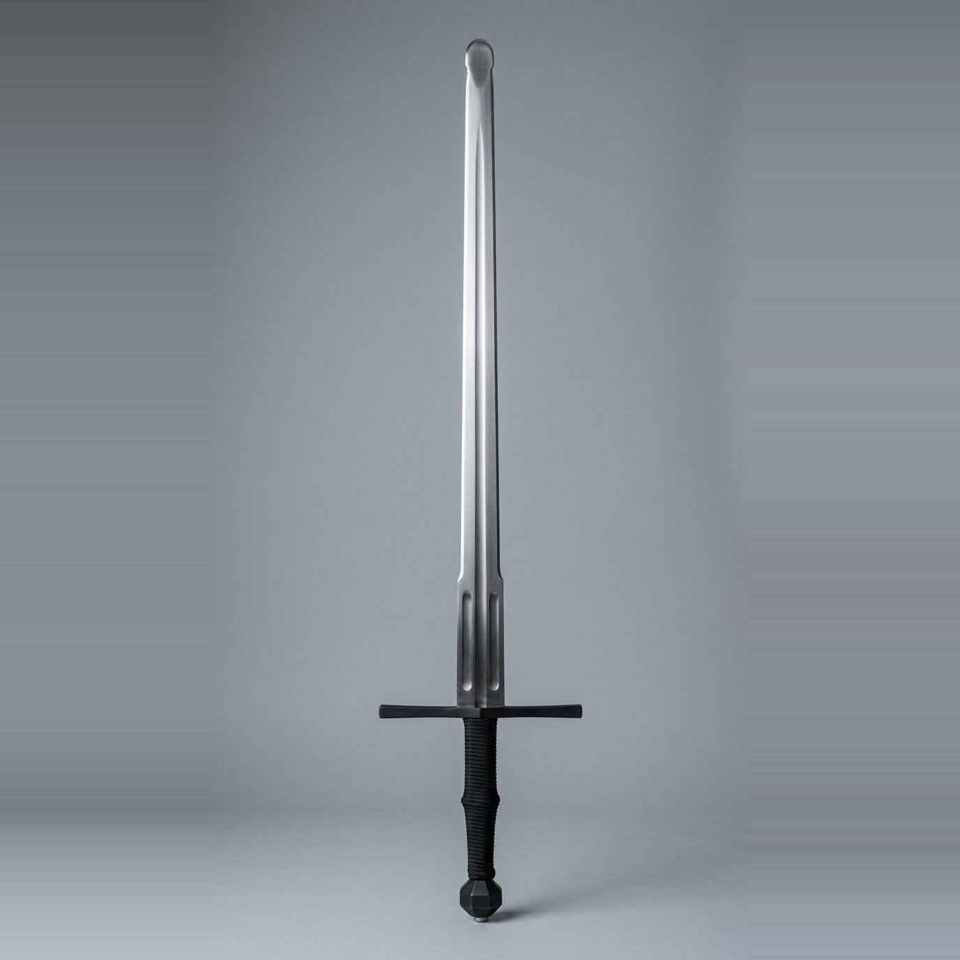 Skarnvik Blackened Longsword Trainer RK-3016 Blackened longsword trainer with twin low fullers, faceted barrel pommel and black cord wrapped grip standing point up against a plain grey studio backdrop