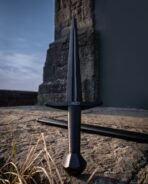 Blackened steel arming sword with a curved guard, knotwork etching and faceted pommel standing on stone, black scabbard behind