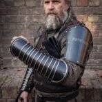 Blackened steel articulated manica on the forearm and rune cut bracer on the upper arm worn by a grey bearded man with a braided beard