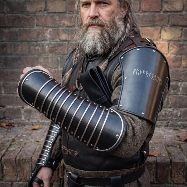 Blackened steel articulated manica on the forearm and rune cut bracer on the upper arm worn by a grey bearded man with a braided beard
