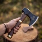 Blackened steel bearded axe head with bright edge and etched hammer symbol, rosewood haft with brass bands in a hand