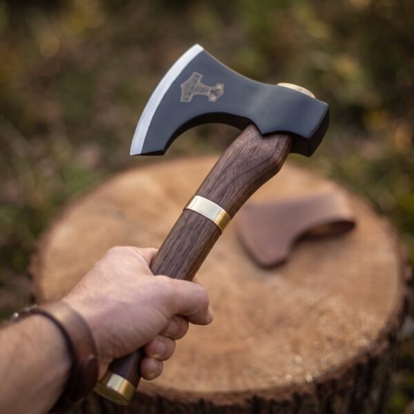 Blackened steel bearded axe with etched hammer emblem, brass collar and rosewood haft held in hand over a tree stump