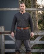 Blackened steel chainmail hauberk with elbow length sleeves worn over grey trousers with a red leather belt by a bearded man at a wooden gate