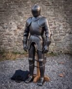 Blackened steel full suit of plate armour on a wooden stand, front three quarter view by a stone wall with padded gambeson
