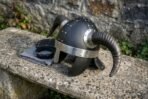 Blackened steel horned helmet with polished horn collars and cheek guards resting on a stone bench in side view