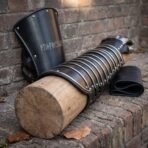 Blackened steel manica of riveted articulated lames with bright edges resting on a wooden log beside an upright rune cut bracer on a brick wall